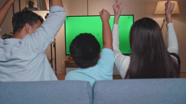 Back View Of Excited Asian Family Watching Tv With Mock Up Green Screen Together Sitting On A Sofa In The Living Room At Home
