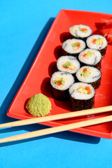 Japanese sushi rolls served on a red plate with pickled ginger and wasabi sauce over colorful blue background.