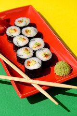 Japanese sushi rolls served on a red plate with pickled ginger and wasabi sauce over green and yellow background.