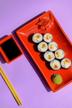 Japanese sushi rolls served on a red plate with pickled ginger and wasabi sauce over colorful purple background.