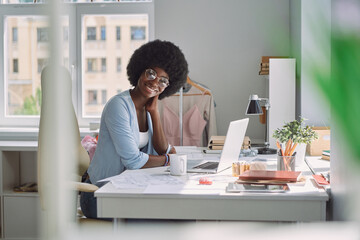 Beautiful young African woman looking at camera and smiling while sitting at her working place in design studio