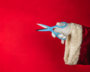 Santa's Hand holding blue scissors, isolated on bright red background with copy space.