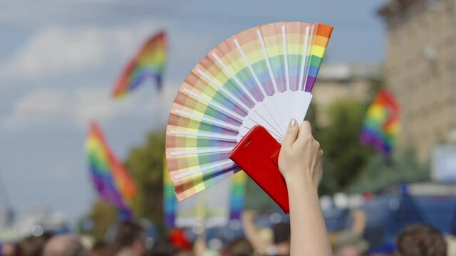 Female hand holding telephone and fan with gay pride colors, flags on blurred background. Partial view of contestants of lgbt parade. Concept of tolerance and equality
