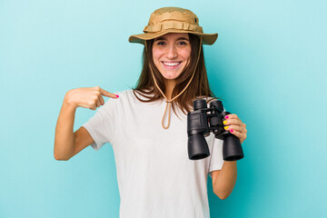 Young caucasian explorer woman holding binoculars isolated on blue background person pointing by hand to a shirt copy space, proud and confident