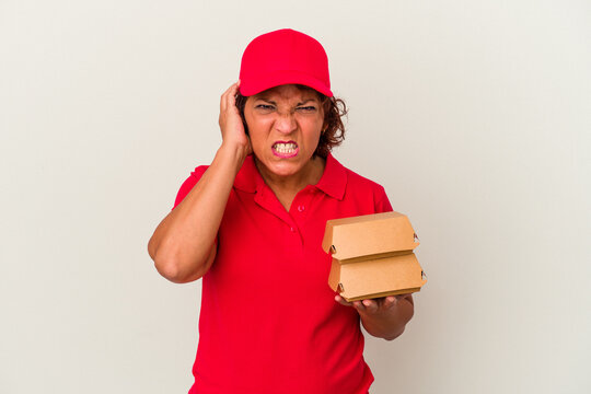Middle age delivery woman taking burguers isolated on white background covering ears with hands.