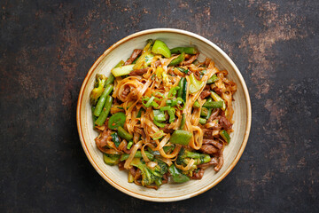 Oriental noodles, top view. Beef fried on a wok with rice noodles, green beans, zucchini, pak choi and green peppers in spicy soy-oyster sauce, in a bowl, on black stony background. Thai cousine dish.