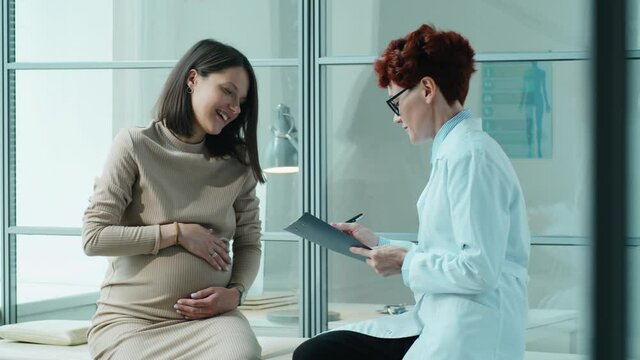 Young pregnant woman sitting on medical couch and speaking with female doctor as she writing down some notes during prenatal checkup in clinic
