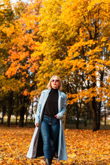 Beauty fashion business woman in stylish clothes with coat and sunglasses walks in autumn park with amazing colored yellow fall leaves outdoors