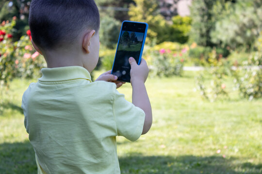 A little boy (kid) takes photos and videos in a park with a phone in blue case. Summertime, green environment.