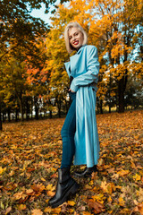 Stylish happy woman with a smile in a trendy autumn blue coat with jeans and fashion fall shoes in an autumn park in nature