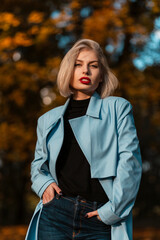 Fashion portrait of a beautiful blonde girl with red lips in a blue coat and a black sweater walks in a park with colored autumn foliage at sunset