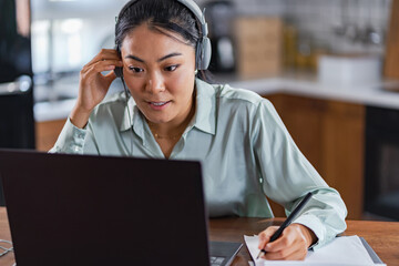 Fototapeta premium Young Asian woman attending online foreign language classes. Sitting in front of laptop computer with headphones listening course and taking notes.