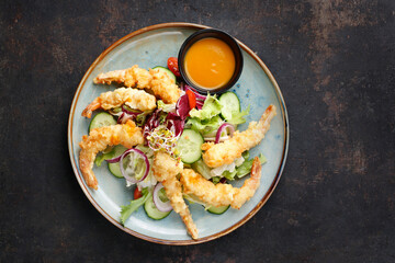 Top view of a salad with prawns in panko, served with oriental sauce based on mango and garlic. Oriental salad mix with fried shrims on a blue plate, on black stony background. Thai cousine.