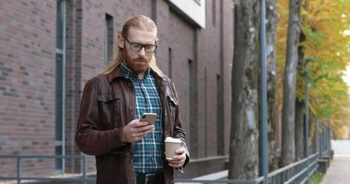 Handsome man walking on the street with smartphone and drinking coffee. Ginger guy wearing glasses with smile on his face messaging at the mobile phone while walking outdoor