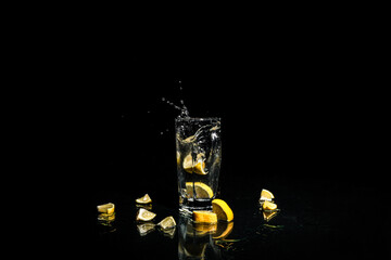 A slice of lemon falls into a glass, black background. Flying water drops