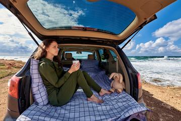A relaxed young woman is resting in the car trunk in front on the ocean..