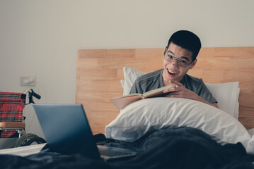 Asian handicapped teenager boy using computer and listening music video in the morning on bed and wheelchair in room, Lifestyle activities of disabled child using technology and remote learning.