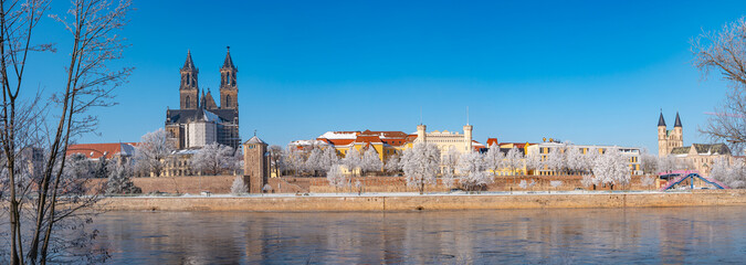 Panoramic view over Magdeburg historical downtown in Winter with icy trees and snow during sunrise in the morning with warm illumination and blue sky, Germany. - obrazy, fototapety, plakaty