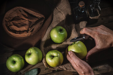 Hands cutting green apple, rustic image with several apples and hat on sackcloth on wooden table.
