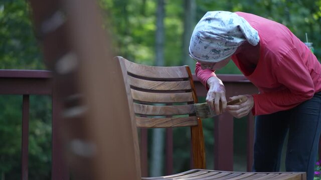 Mature woman applying varnish to teak table furniture in garden area at home.