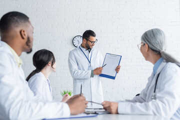 Fototapeta premium young doctor pointing at medical card on clipboard near blurred multicultural colleagues