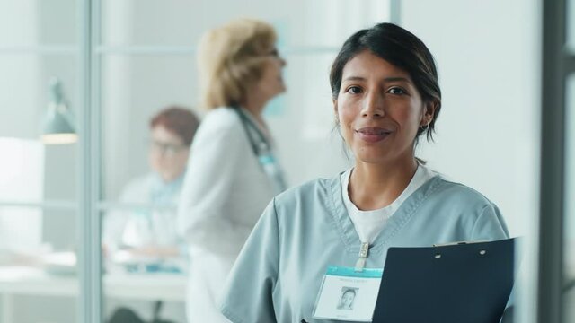 Portrait of cheerful Hispanic nurse in medical uniform holding clipboard, looking at camera and smiling while standing in clinic