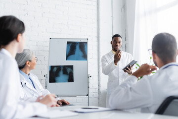 african american doctor gesturing while talking to blurred colleagues near flip chart with lungs x-rays