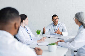 doctor in eyeglasses gesturing while talking to blurred multiethnic colleagues
