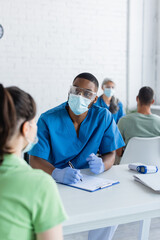 african american doctor with clipboard and pen talking to woman in medical mask, vaccination concept