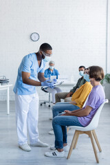 african american doctor in medical mask holding clipboard while talking to patients in vaccination center
