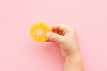 Baby hand holding ring shape of yellow candied fruit jelly on light pink table background. Pastel...