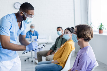 african american doctor in medical mask writing on clipboard near patients in vaccination center