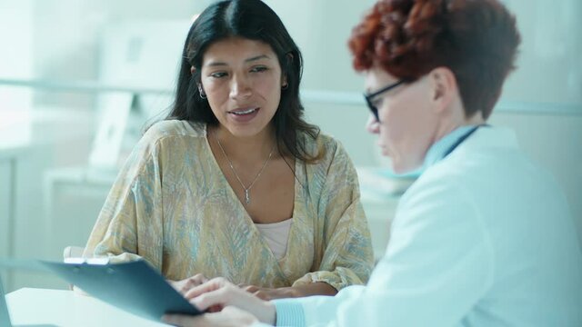 Beautiful Hispanic woman smiling and listening to Caucasian female doctor as she explaining her something on clipboard during health consultation in medical office