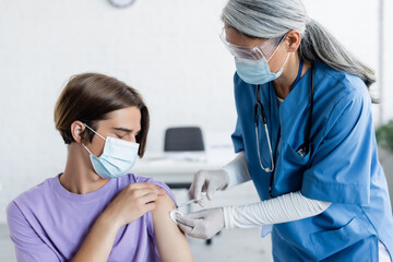 mature asian doctor in medical mask and goggles giving vaccine injection to young patient