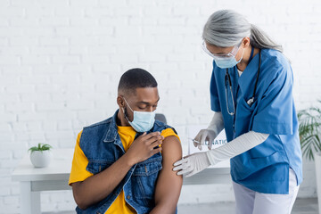 african american man in medical mask near asian nurse holding syringe with vaccine