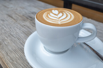 Selected focus and close up view of a white ceramic cup of cappuccino on top with beautiful delicate latte art with flower and heart pattern, on wooden outdoor counter of coffee shop.