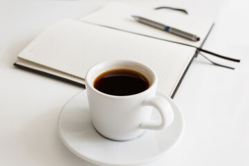 The concept of a workplace with coffee in a neat white mug on a saucer and an open notebook with a pen on a modern white office desk. Break for a delicious and hot coffee. Minimalism. Side view