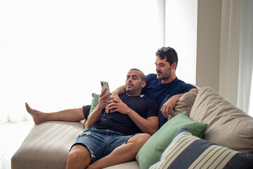 couple of gay men in love, relaxing on the sofa in their living room, fiddling with their cell phone, researching the new summer vacation

