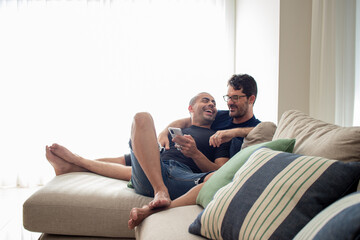 couple of gay men in love, relaxing on the sofa in their living room, fiddling with their cell phone, researching the new summer vacation

