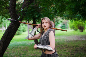 Portrait of a medieval, fantasy warrior girl, in chain mail armor, armed with a spear. Posing against the background of the forest.