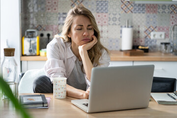 Unhappy woman of middle age search for new job unemployed during covid-19 quarantine. Female look at laptop screen with vacancies sitting unpleased on home kitchen in pajama. Unemployment concept - obrazy, fototapety, plakaty