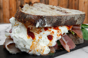 Gourmet sandwich. Mediterranean flavors. Closeup view of an Italian burrata cheese, blue cheese, arugula and cured ham sandwich in a black dish on the wooden table.
