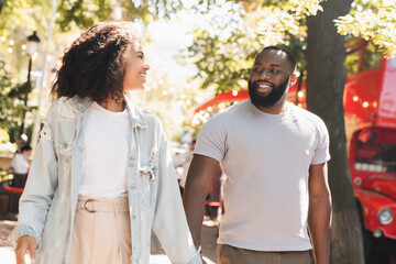 Naklejka premium Cheerful young african couple walking together on romantic date outdoors in city park. Love and relationship, bonding concept
