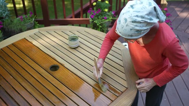 Overhead view of mature woman applying varnish to teak table furniture in garden area at home.