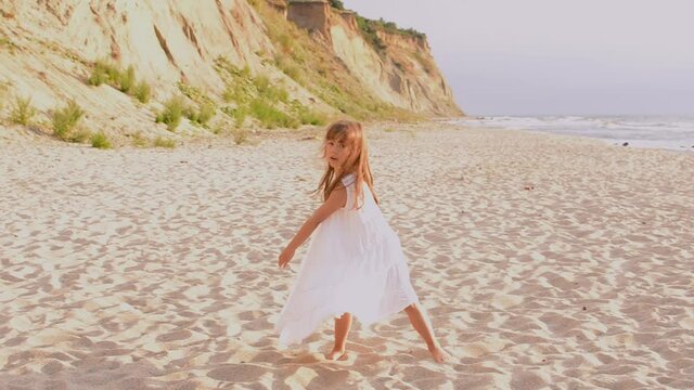 Adorable happy little girl in white dress on white beach at sunset.