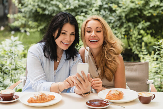 Cheerful smiling laughing two caucasian middle-aged women best friends businesswomen showing news photos on social media using smart phone cellphone in cafe during breakfast
