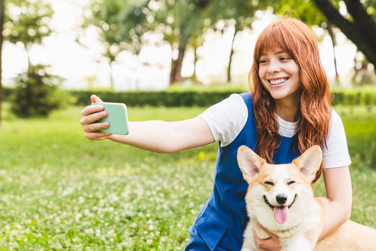 Beautiful caucasian young woman girl teenager with ginger hair taking selfie photo with dog pet welsh corgi on smart phone, having video call conversation online while walking in park