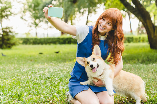 Caucasian red-haired woman young teenager girl taking selfie photo together with her welsh corgi dog pet, having online video call conversation on smart phone in park.