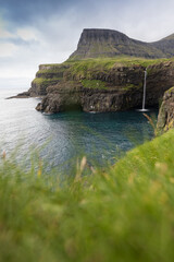 Famous Gasadalur waterfall from viewpoint during a sunny summer day with mountain peaks and calm sea. Amazing cliffs on the Faroe islands, Denmark, Europe. A view of high peaks of mountains.