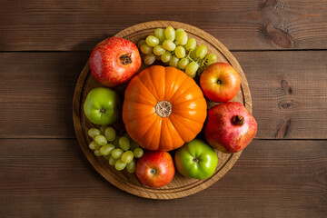 Autumn frame still life from ripe fruits orange pumpkin, apples, grape and garnet. Fall harvest on round wooden cutting board and brown background flat lay.